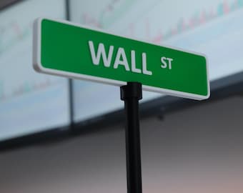 May include: A green and white "WALL ST" street sign stands in front of two computer monitors displaying financial charts. The sign is mounted on a black post, suggesting a theme related to finance or the stock market.