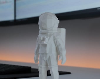 May include: A white, low-poly geometric figurine of an astronaut standing upright. The figure features a faceted, angular design with a smooth matte finish, a rounded helmet, and a rectangular life support backpack attached to the back of the suit.