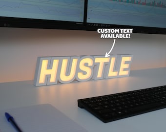May include: A white, illuminated "HUSTLE" sign with a warm yellow glow. The sign is on a white desk, next to a keyboard and notepad. The text "CUSTOM TEXT AVAILABLE!" is also visible.