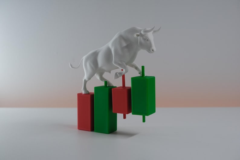 Op de afbeelding: Een wit stierenbeeldje dat bovenop een reeks driedimensionale rode en groene rechthoekige balken van een financiële candlestick-grafiek staat. De stier is gepositioneerd met de voorpoten omhoog, balancerend op de gekleurde blokken.