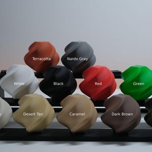 May include: A collection of eleven twisted, spherical objects arranged on a black tiered display stand. Each object is a different solid color labeled as Terracotta, Nardo Gray, White, Black, Red, Green, Bone White, Desert Tan, Caramel, Dark Brown, and Dark Chocolate.