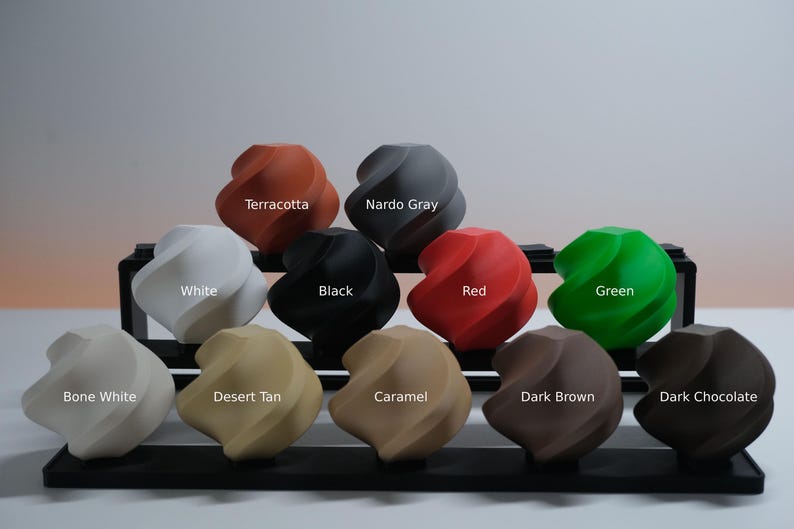 May include: A collection of eleven twisted, spherical objects arranged on a black tiered display stand. Each object is a different solid color labeled as Terracotta, Nardo Gray, White, Black, Red, Green, Bone White, Desert Tan, Caramel, Dark Brown, and Dark Chocolate.