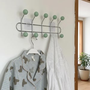 Puede incluir: Un perchero de pared con seis ganchos, cada uno rematado con una esfera verde claro. Una camisa de pijama a cuadros azul claro y blanco con motivos de osos cuelga de una percha blanca. Una camisa blanca cuelga a la derecha.