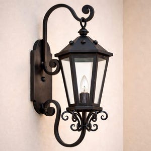 May include: Black outdoor wall-mounted lantern with clear glass panels. The lantern has a decorative scrolled arm and bracket. The light fixture features a classic design with an Edison-style bulb. The lantern is mounted on a cream wall.