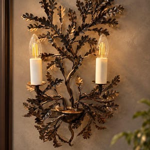 May include: A wall sconce featuring a dark bronze-toned metal frame shaped like a branching oak tree with numerous leaves. Two vertical white candle-style light sockets are positioned on the branches, each holding a clear filament light bulb.