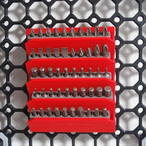 May include: A red organizer with multiple rows of silver and gray screwdriver bits. The organizer is mounted on a black, geometric patterned surface. The bits are of various shapes and sizes, suitable for different screw types.