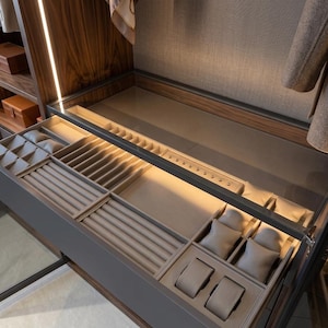 May include: A gray, open drawer with compartments for jewelry and watches. The drawer is lined with beige fabric and features various sections for rings, bracelets, and other accessories. The drawer is part of a larger wooden cabinet.