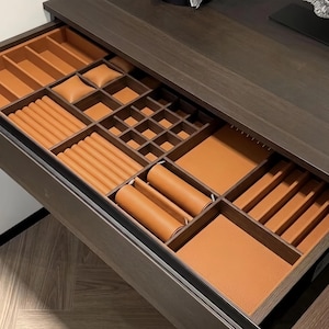 May include: A dark wood dresser drawer featuring a custom tan leather-lined jewelry organizer insert. The interior includes multiple compartments, including ring rolls, watch pillows, square dividers, and open storage sections for organizing accessories.