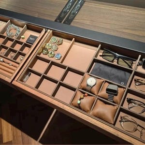May include: A wooden jewelry and accessories organizer with multiple compartments. It contains jewelry, watches, glasses, and other accessories. The organizer is brown with tan-colored compartments and brown leather watch pillows.