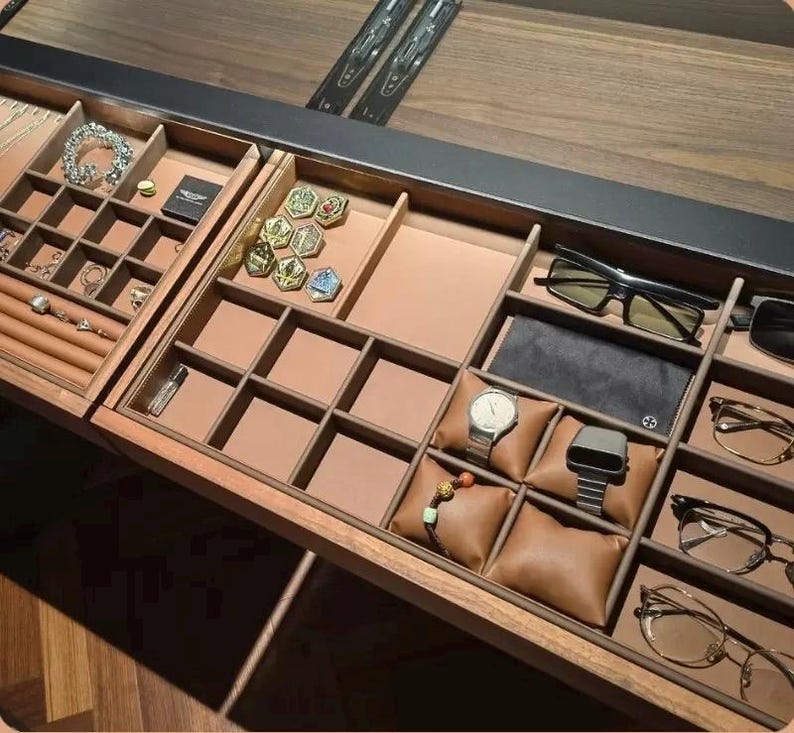 May include: A wooden jewelry and accessories organizer with multiple compartments. It contains jewelry, watches, glasses, and other accessories. The organizer is brown with tan-colored compartments and brown leather watch pillows.