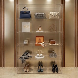 May include: A clear acrylic display case filled with luxury items. The shelves hold handbags, a book with "DIOR: COUTURE" on the cover, perfume, jewelry, and shoes. The items are in various colors, including blue, gray, white, and beige.