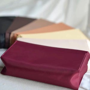Peut inclure: Plusieurs pochettes rectangulaires de différentes couleurs, dont bordeaux, noir, marron, beige et crème. Les pochettes sont disposées en éventail, avec la pochette bordeaux au premier plan. Elles semblent être faites d'un matériau lisse, peut-être satiné.