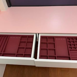 May include: Open white drawer with two burgundy jewelry organizer trays. The trays have various compartments for rings, necklaces, and other jewelry. The drawer is part of a pink and white vanity.