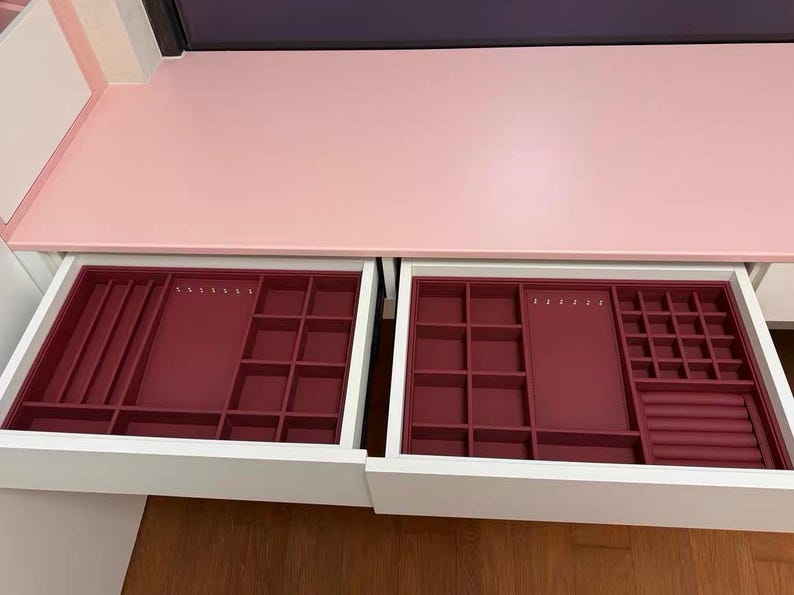 May include: Open white drawer with two burgundy jewelry organizer trays. The trays have various compartments for rings, necklaces, and other jewelry. The drawer is part of a pink and white vanity.
