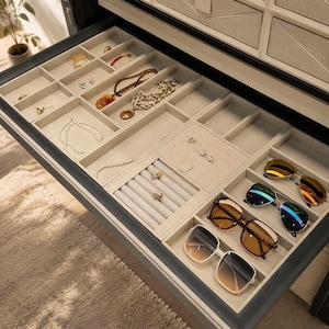 May include: An open jewelry drawer organizer with a beige velvet interior featuring multiple compartments. It contains various gold-toned necklaces, chains, rings, and four pairs of sunglasses with different lens tints arranged in side slots.