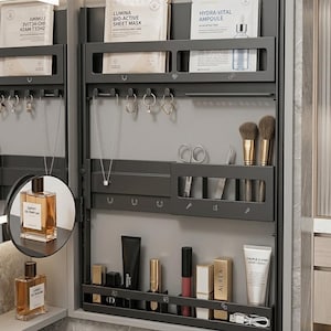 Puede incluir: Un armario de almacenamiento gris oscuro montado en la pared con múltiples estantes y ganchos. Contiene productos para el cuidado de la piel, maquillaje, joyas y frascos de perfume. El armario tiene un espejo incorporado con una vista ampliada de un frasco de perfume.