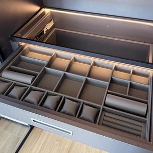 May include: Open gray jewelry drawer with multiple compartments and watch pillows. The drawer is illuminated by a strip of light at the top. The drawer is part of a larger gray cabinet.