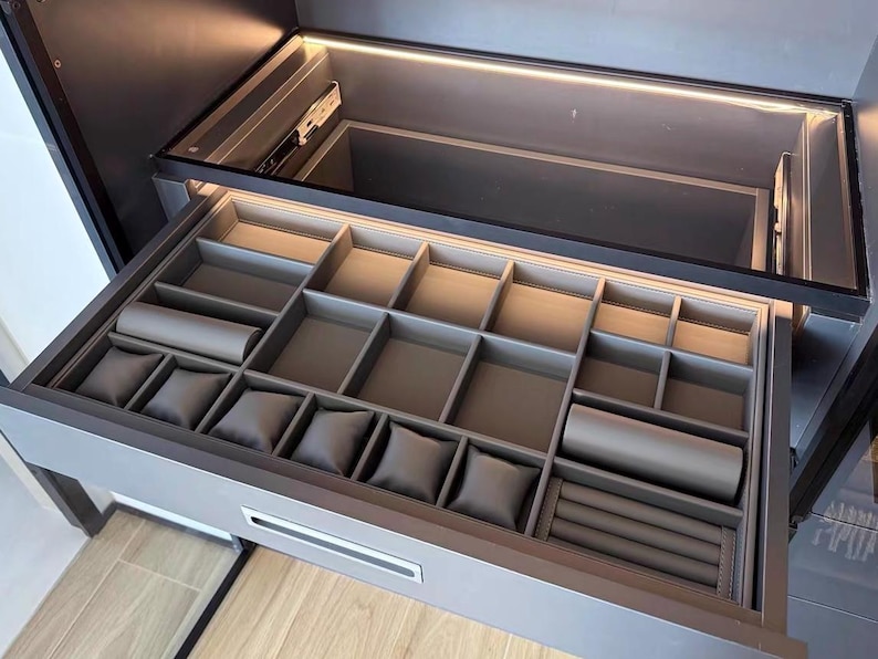 May include: Open gray jewelry drawer with multiple compartments and watch pillows. The drawer is illuminated by a strip of light at the top. The drawer is part of a larger gray cabinet.