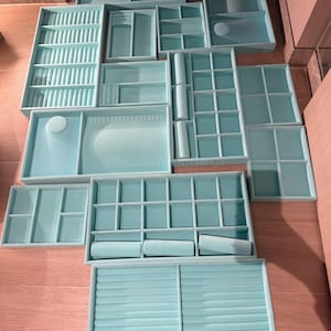 May include: A collection of light blue jewelry organizer trays in various shapes and sizes. The trays include compartments, ring holders, and roll-style storage. The organizers are arranged on a wooden surface.
