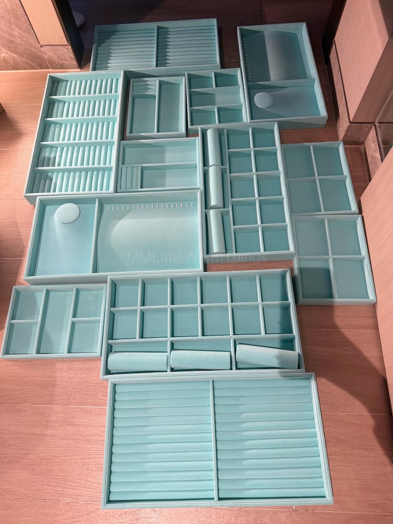May include: A collection of light blue jewelry organizer trays in various shapes and sizes. The trays include compartments, ring holders, and roll-style storage. The organizers are arranged on a wooden surface.