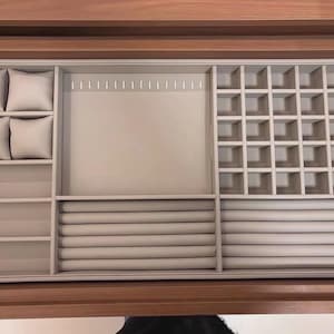 May include: A beige velvet jewelry drawer organizer insert featuring four square watch pillows, a large open compartment, a grid section with thirty-two small square slots, and two sections containing a total of ten horizontal ring display rolls.