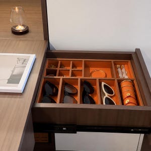 May include: A dark wood drawer organizer with orange velvet lining, featuring multiple compartments containing four pairs of sunglasses, a gold bangle, various small jewelry pieces, and a cylindrical jewelry roll with gold bracelets.