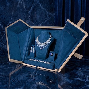 Puede incluir: Un lujoso conjunto de joyas exhibido en una caja de terciopelo azul. El conjunto incluye un collar, pendientes, un anillo y una pulsera, todos con piedras preciosas plateadas y azules. La caja tiene un borde dorado y está abierta.