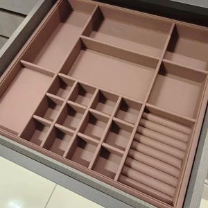 May include: A gray wooden drawer with a pink interior jewelry organizer. The organizer has various compartments of different sizes, including small square sections and long, narrow slots for rings. The interior is a solid, muted pink color.