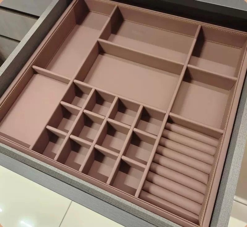 May include: A gray wooden drawer with a pink interior jewelry organizer. The organizer has various compartments of different sizes, including small square sections and long, narrow slots for rings. The interior is a solid, muted pink color.
