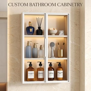 以下が含まれることがあります： 白い壁掛け式のバスルームキャビネットで、2つのドア、照明付きの棚、上部に「CUSTOM BATHROOM CABINETRY」の文字があります。キャビネットには、ボトル、ディフューザー、歯ブラシホルダーなど、さまざまな化粧品が展示されています。