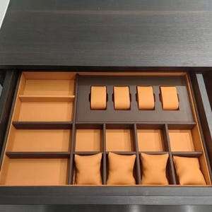May include: A dark wood drawer with an open interior. The drawer is divided into compartments with orange and brown accents. The compartments include watch holders and small pillow-like cushions, designed for storing jewelry and watches.