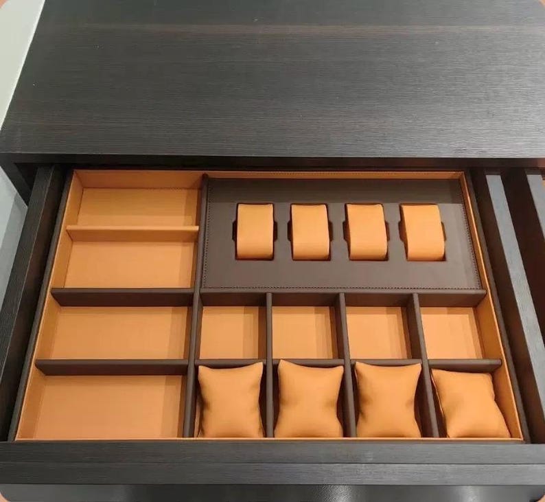 May include: A dark wood drawer with an open interior. The drawer is divided into compartments with orange and brown accents. The compartments include watch holders and small pillow-like cushions, designed for storing jewelry and watches.