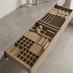 May include: Open wooden jewelry organizer drawer with multiple compartments and beige velvet lining. The drawer is designed to hold various jewelry items, including rings, bracelets, and watches. The organizer is set on a gray countertop.