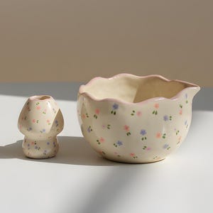 May include: A ceramic mushroom-shaped vase and a bowl with a wavy rim. Both are off-white and decorated with small pink, blue, and green floral patterns. The bowl has a pink rim.