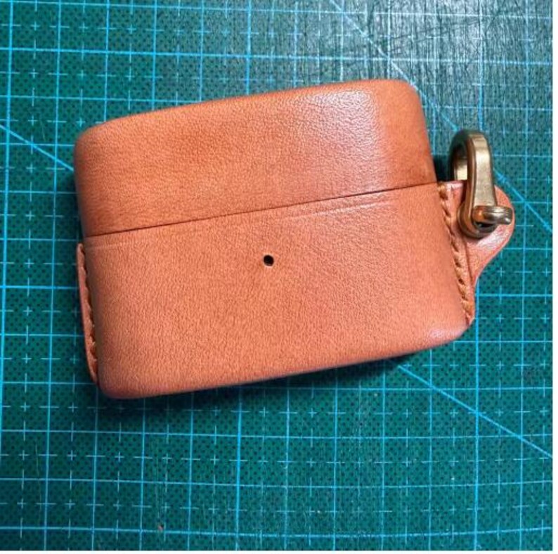 K&ouml;nnte beinhalten: Eine hellbraune Schutzh&uuml;lle aus Leder f&uuml;r kabellose Ohrh&ouml;rer mit glatter Textur, sichtbaren N&auml;hten an der Seite, einer kleinen mittigen kreisf&ouml;rmigen Aussparung und einem goldfarbenen Karabinerhaken an einer Lederlasche auf der rechten Seite.
