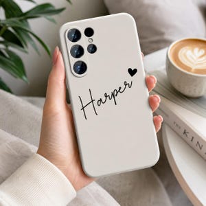 以下が含まれることがあります： マットなオフホワイトのスマートフォンケース。黒い筆記体で「Harper」という名前と、小さな黒いハートのアイコンが描かれています。ケースの左上隅には、マルチレンズカメラシステム用の正確な切り抜きが施されています。