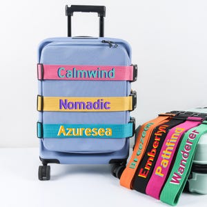 Puede incluir: Una maleta rodante azul claro con tres correas de colores que dicen "Calmwind", "Nomadic" y "Azuresea". Varias correas de equipaje con nombres se muestran a la derecha, incluyendo "Breezely", "Emberlyn", "Pathfind" y "Wanderer".