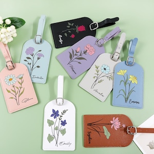 Könnte beinhalten: Eine Sammlung von Gepäckanhängern in verschiedenen Farben, jeweils mit floralem Design und Namen. Die Anhänger haben eine rechteckige Form mit abgerundeten Kanten und einem Riemen zur Befestigung. Farben sind u.a. Rosa, Schwarz, Weiß, Lila und Braun.