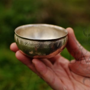 Könnte beinhalten: Eine kleine, silberfarbene Schale mit einem dekorativen geätzten Muster am Rand. Die Schale wird in einer Hand vor einem verschwommenen grünen Hintergrund gehalten, was auf eine Umgebung im Freien hindeutet. Das Innere der Schale reflektiert Licht.