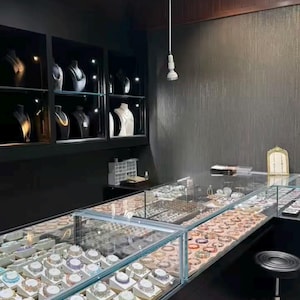 May include: Jewelry store interior with display cases filled with necklaces, bracelets, and rings. The necklaces are displayed on mannequins in glass-fronted cabinets. The store has a modern design with black walls and bright lighting.