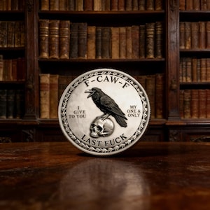 May include: A silver coin with a detailed design featuring a crow perched atop a skull. The coin has the text "F-CAW-F", "I GIVE TO YOU", "MY ONE & ONLY", and "LAST FUCK" around the edges. The background is a blurred image of a bookshelf.