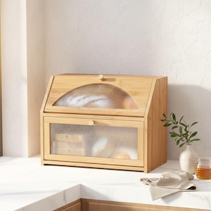 May include: A bamboo bread box with a clear, textured glass door and a curved top. The box has a hinged lid and a lower compartment, both with glass panels. The bread box is sitting on a white countertop, with a vase and a jar of honey nearby.