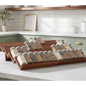 May include: A tiered, dark wood spice rack filled with glass jars of various spices. The rack is on a white countertop in a kitchen with green cabinets and a framed picture.