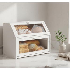May include: A white wooden bread box with a clear glass front and top, filled with various types of bread. The box has a hinged top and a lower door, both with small silver knobs. The bread box is on a white countertop.