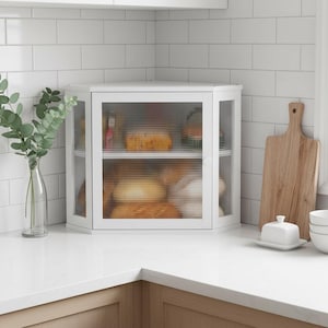 May include: White corner cabinet with two shelves and ribbed glass doors. The cabinet is filled with various baked goods. A wooden cutting board, white bowls, and a butter dish are on the countertop. A vase with greenery is on the left.