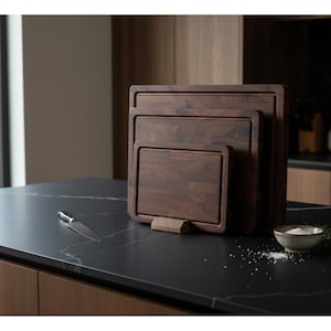 May include: A set of three dark brown wooden cutting boards of varying sizes, stacked together. A small knife rests on the black countertop, along with a small bowl of salt and a sprig of rosemary. The boards are made of a dark wood.