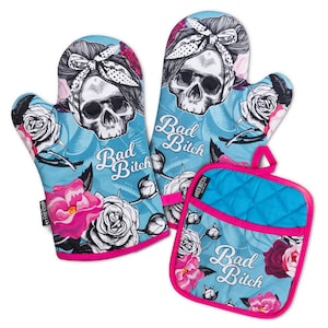 Könnte beinhalten: Zwei Ofenhandschuhe und ein quadratischer Topflappen in Hellblau mit einem Grafikdruck eines Schädels mit gepunktetem Stirnband, umgeben von rosa und weißen Rosen. Der Text Bad Bitch ist in weißer Schreibschrift auf jedem Teil aufgedruckt.