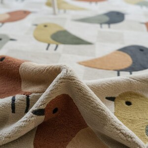 May include: A close-up of a soft, beige blanket with a repeating pattern of colorful birds. The birds feature shades of brown, yellow, green, and blue, set against a checkered background. The blanket's texture is visible in the folded corner.