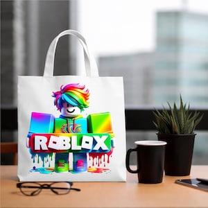 May include: White tote bag with a colourful Roblox character design. The character has rainbow hair and the word "ROBLOX" in a vibrant, dripping paint style. The bag has white handles. The bag is on a wooden table.
