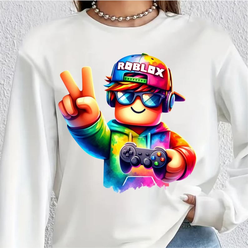 May include: White sweatshirt with a colourful Roblox character giving a peace sign. The character wears a rainbow hat with the word "ROBLOX", sunglasses, and headphones, holding a game controller. The design is bright and striking.